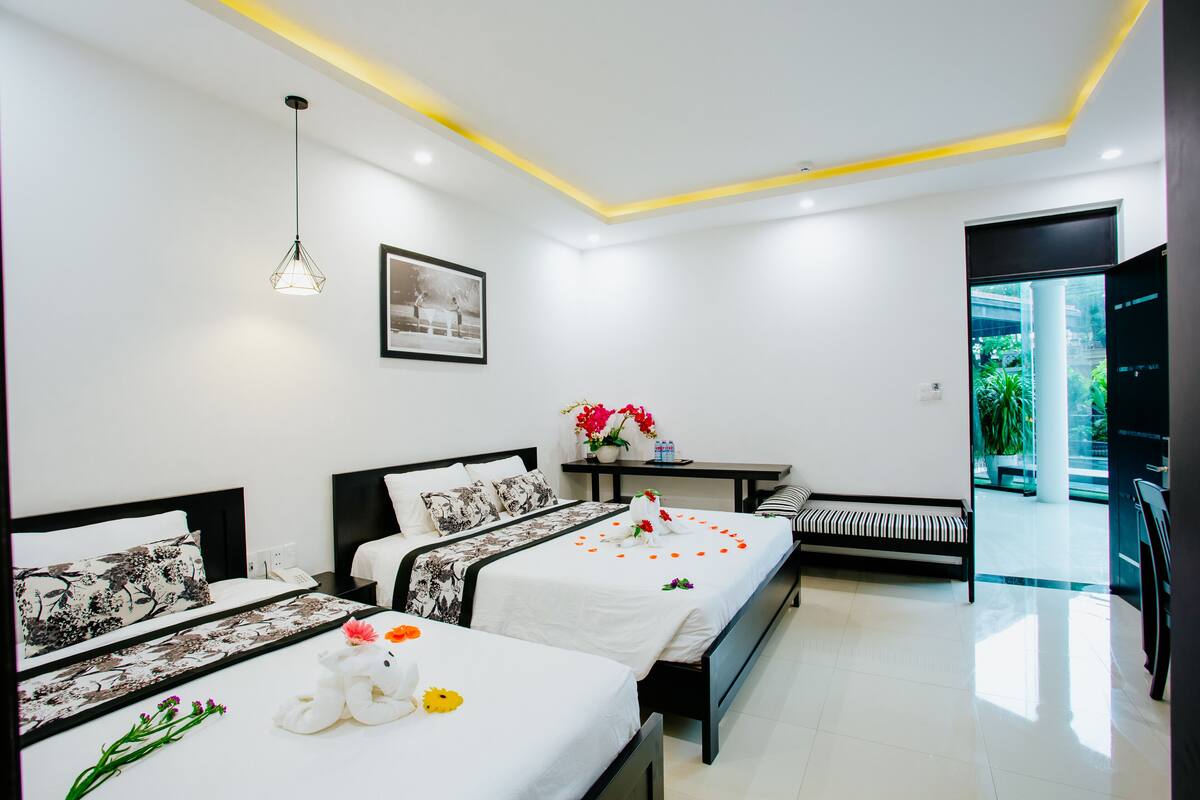 A modern hotel room with two beds, a bench, a small table with flowers, and a view of greenery outside.