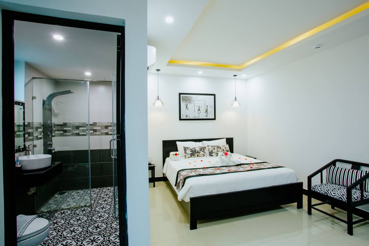 A modern hotel room with a large bed, a separate bathroom with a glass shower enclosure, a small bedside table, and a chair with a patterned cushion.