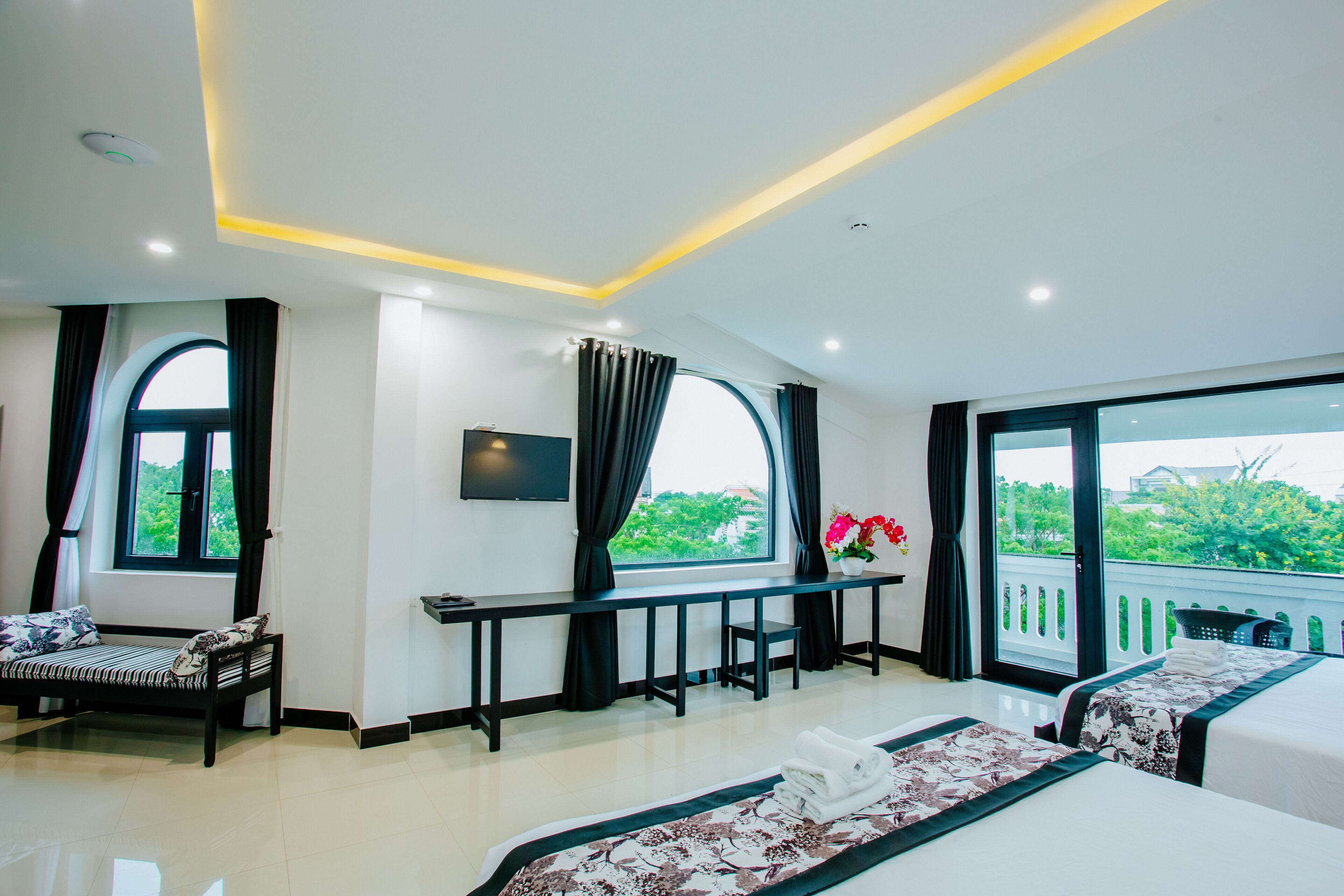 A hotel room with a bed, a desk, a chair, a TV, and a balcony with a view of greenery.