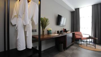 A modern hotel room with a wooden desk, a TV mounted on the wall, a seating area with a chair, and a large window with curtains.