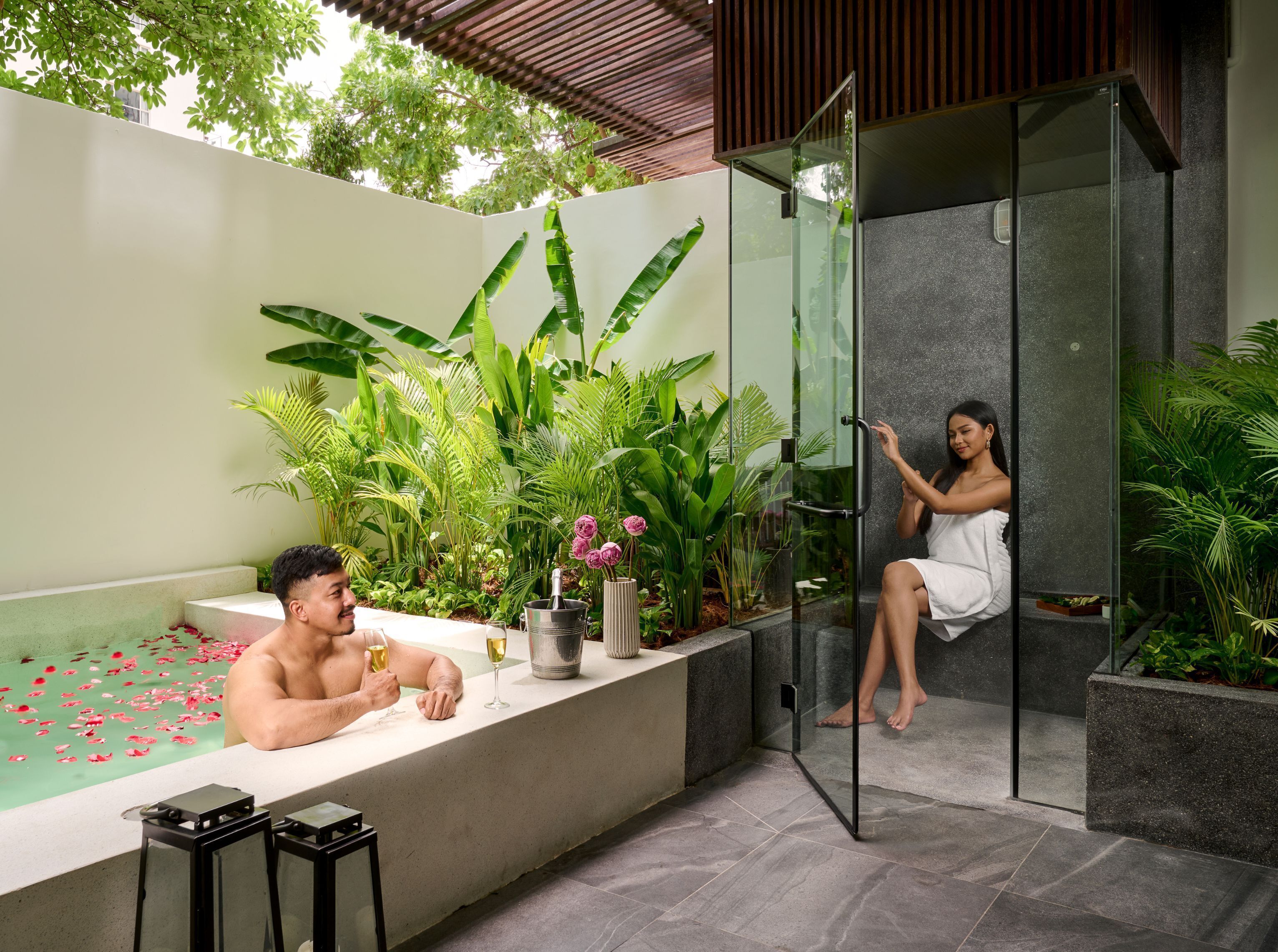 Un moderno espacio de spa con ducha envuelta en vidrio, una bañera con pétalos de rosa y abundante follaje.