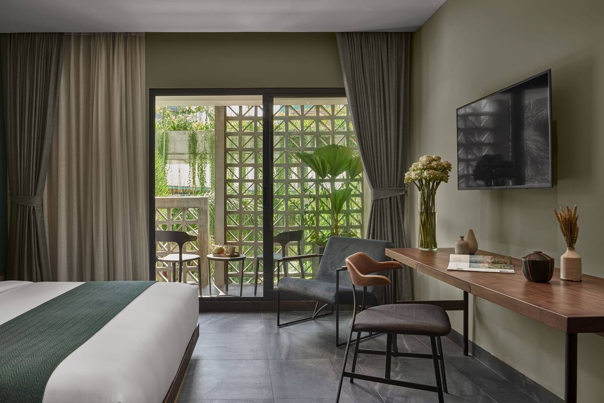 A hotel room with a bed, a desk, two chairs, a table, and a large window with a view of greenery.