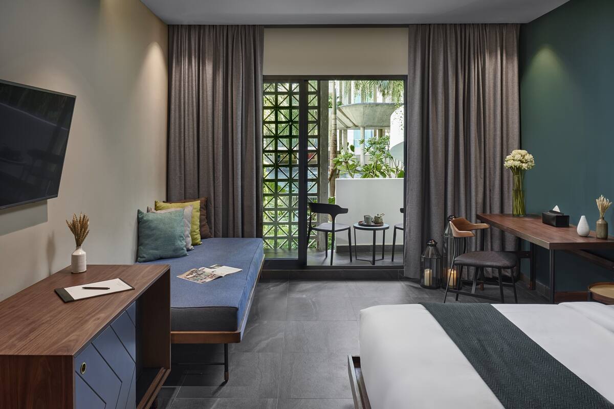 A modern hotel room with a bed, a desk, a chair, a TV, and a balcony with a view of greenery.
