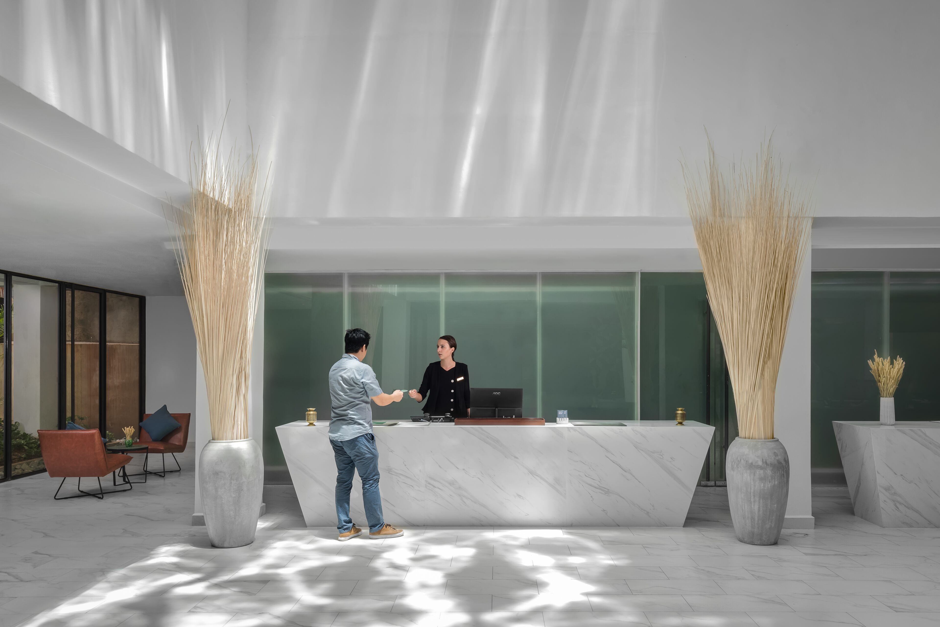 Un área de recepción moderna con mostrador de mármol, dos jarrones altos con pastos y un espacio de descanso con sillas y una mesa.