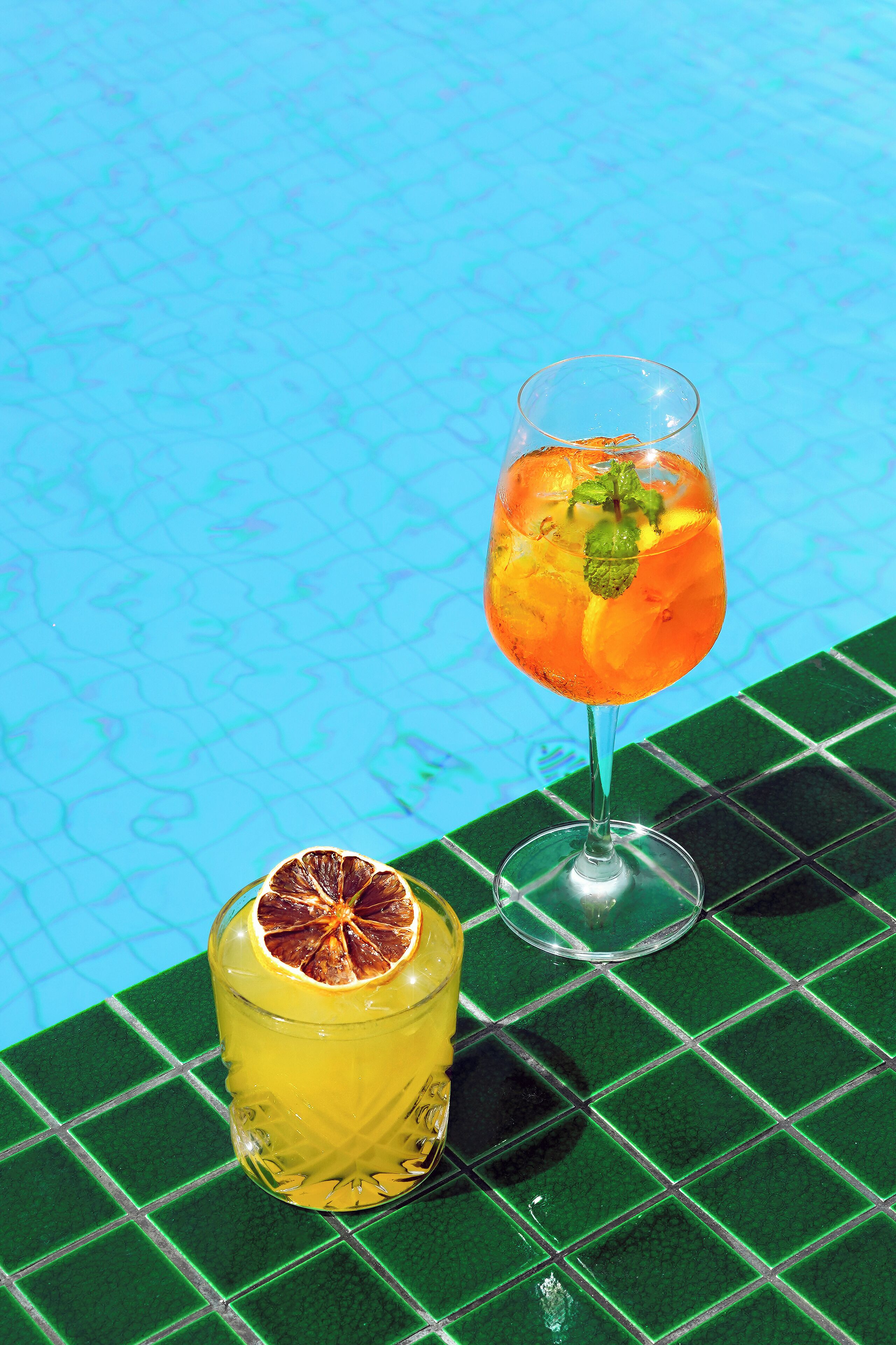 Two glasses of beverages on a tiled surface by a pool.