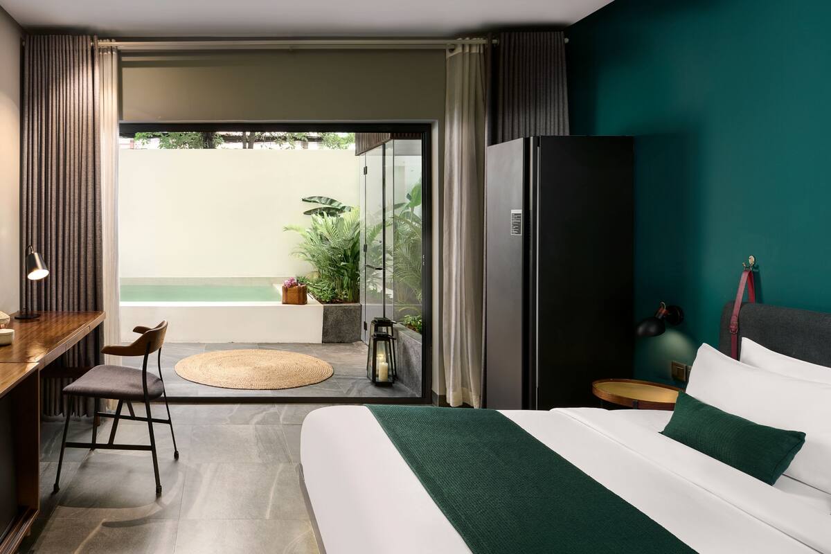A hotel room with a bed, a desk, a chair, a refrigerator, and a view of a pool.