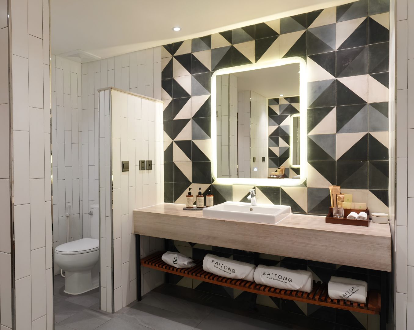 Un baño moderno con una pared de diseño geométrico, un espejo grande, un lavamanos y artículos de aseo ordenados.