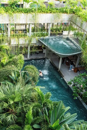 Una piscina interior rodeada de exuberante vegetación, con una persona nadando y otra relajándose junto a la piscina.