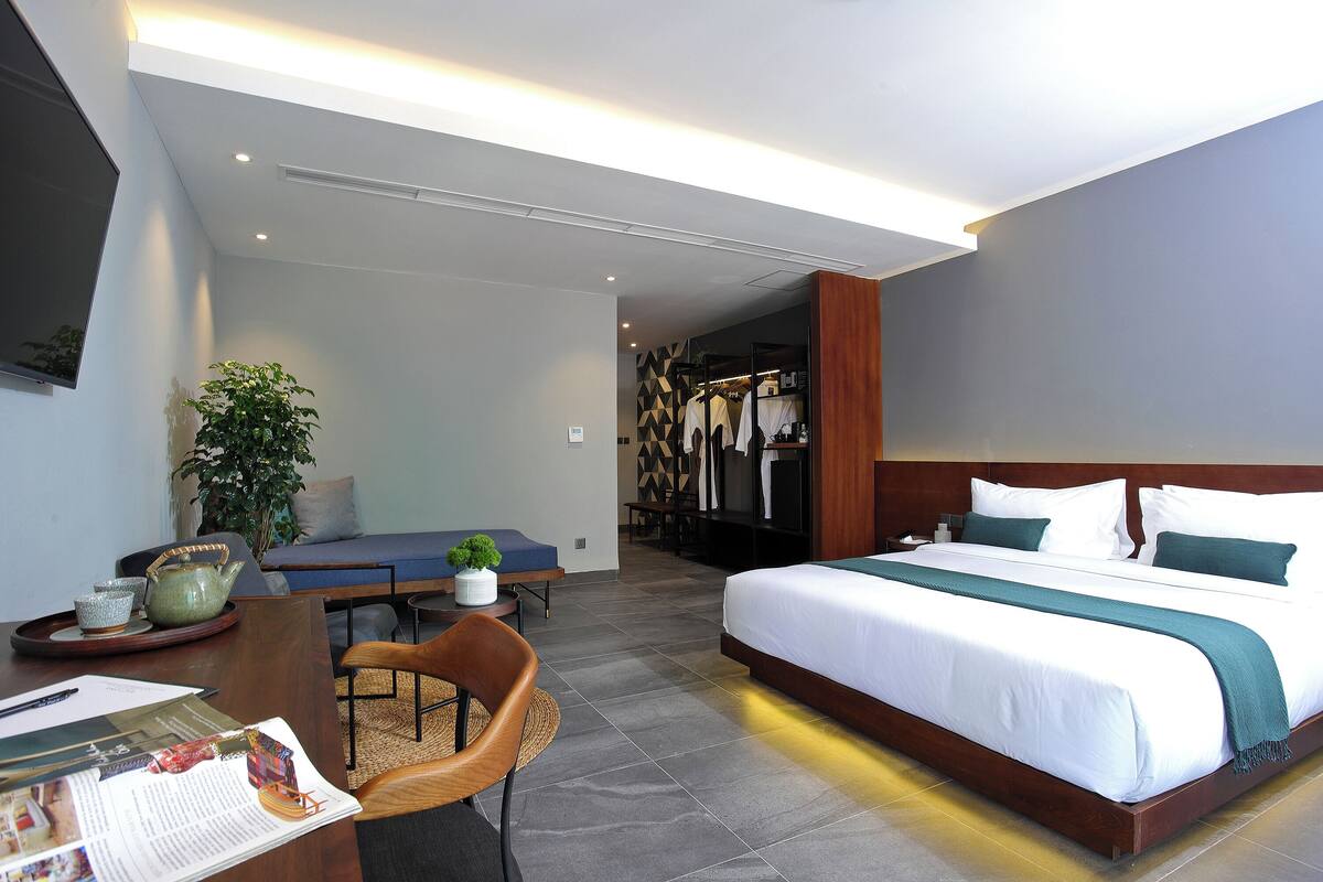 A modern hotel room with a large bed, a desk with a chair, a small table with a plant, and a seating area with a sofa.
