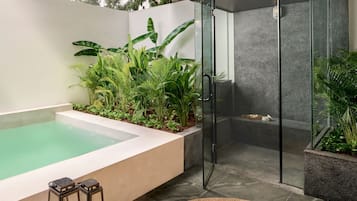 A modern outdoor bathroom with a glass-enclosed shower, a small pool, and lush greenery.