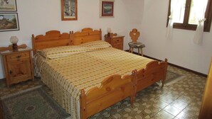 Een slaapkamer met een houten bed, nachtkastje, stoel, klein tafeltje en een raam met gordijnen.