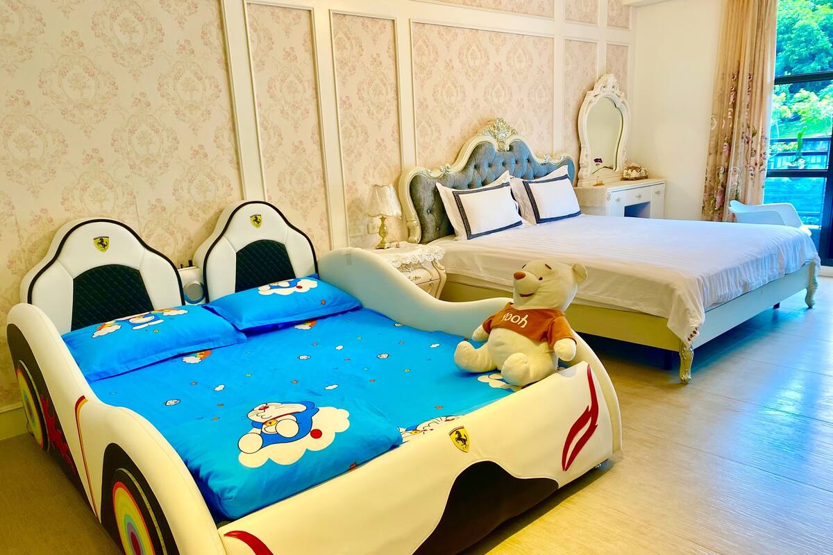 Ein Schlafzimmer mit einem Bett, zwei Stühlen, einem Teddybären, einem Schminktisch und einem Fenster mit Vorhängen.