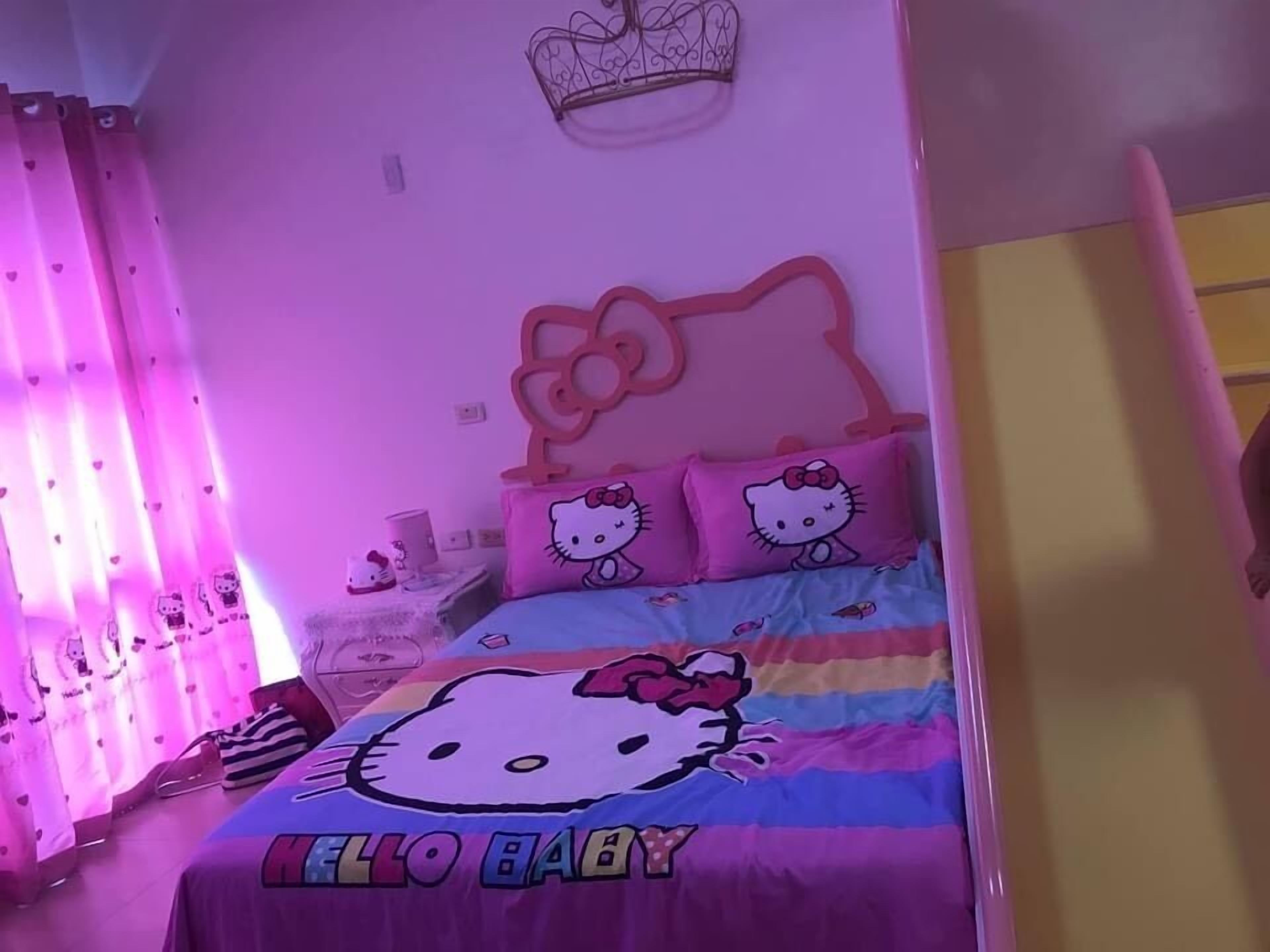 A room with a bed featuring Hello Kitty bedding, pink curtains with Hello Kitty prints, and a Hello Kitty crown on the wall.