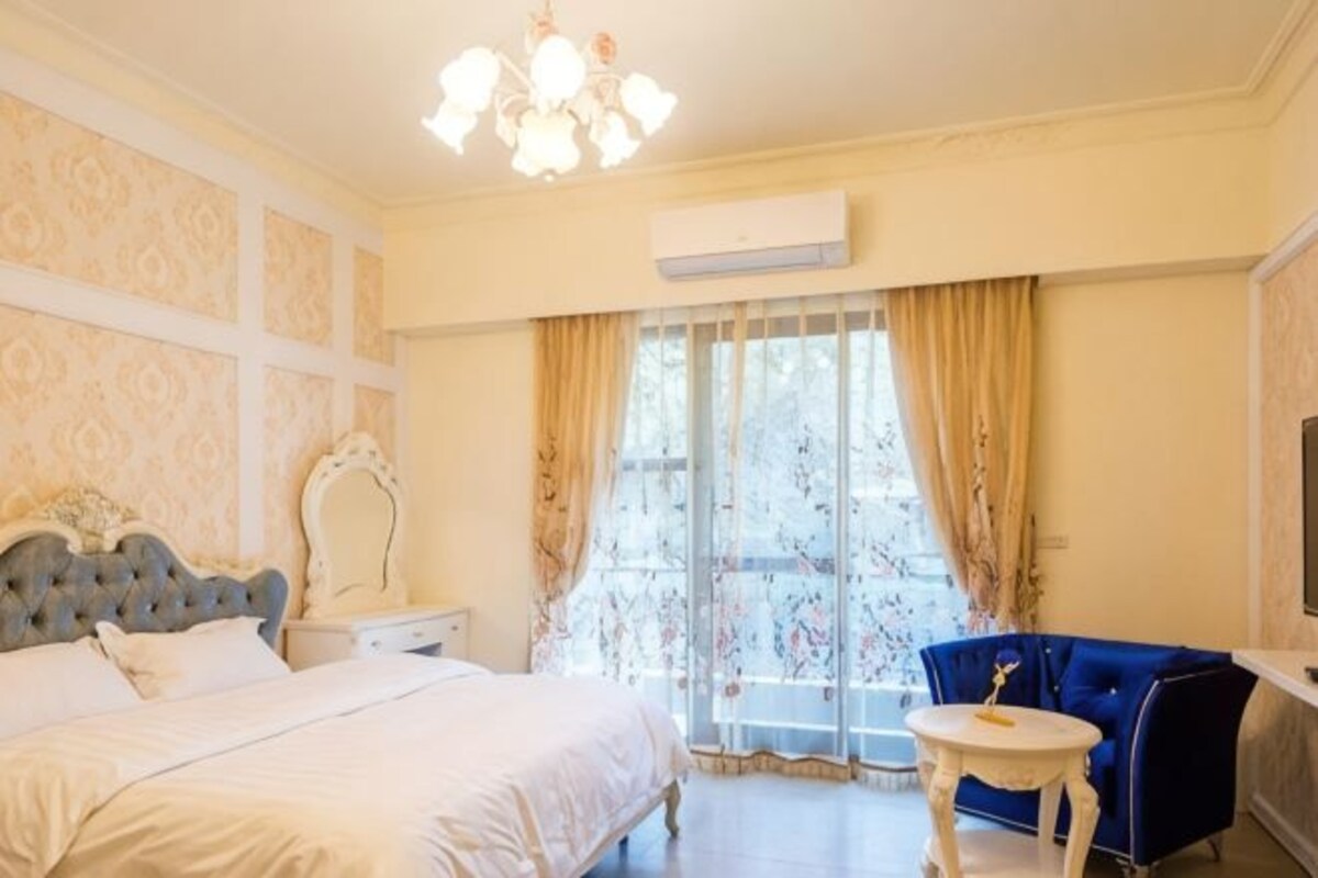 A bedroom with a bed, a vanity, a blue armchair, a small table, and a large window with curtains.