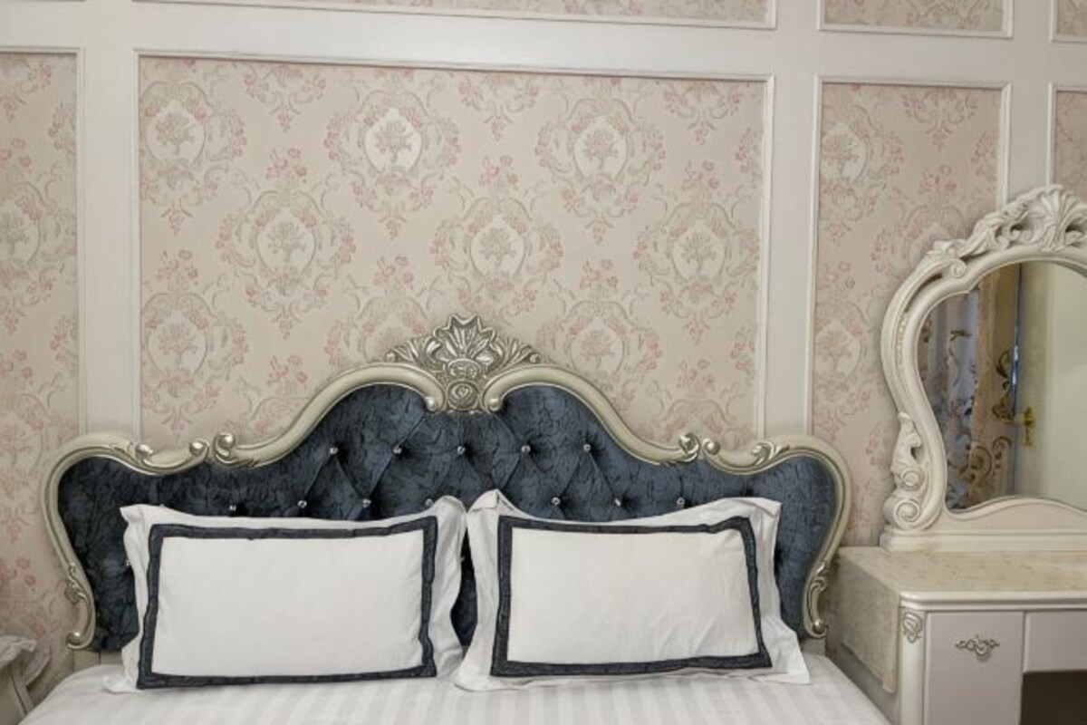 Una cama con cabecera capitonada, dos almohadas, un aparador con espejo y un papel tapiz estampado.