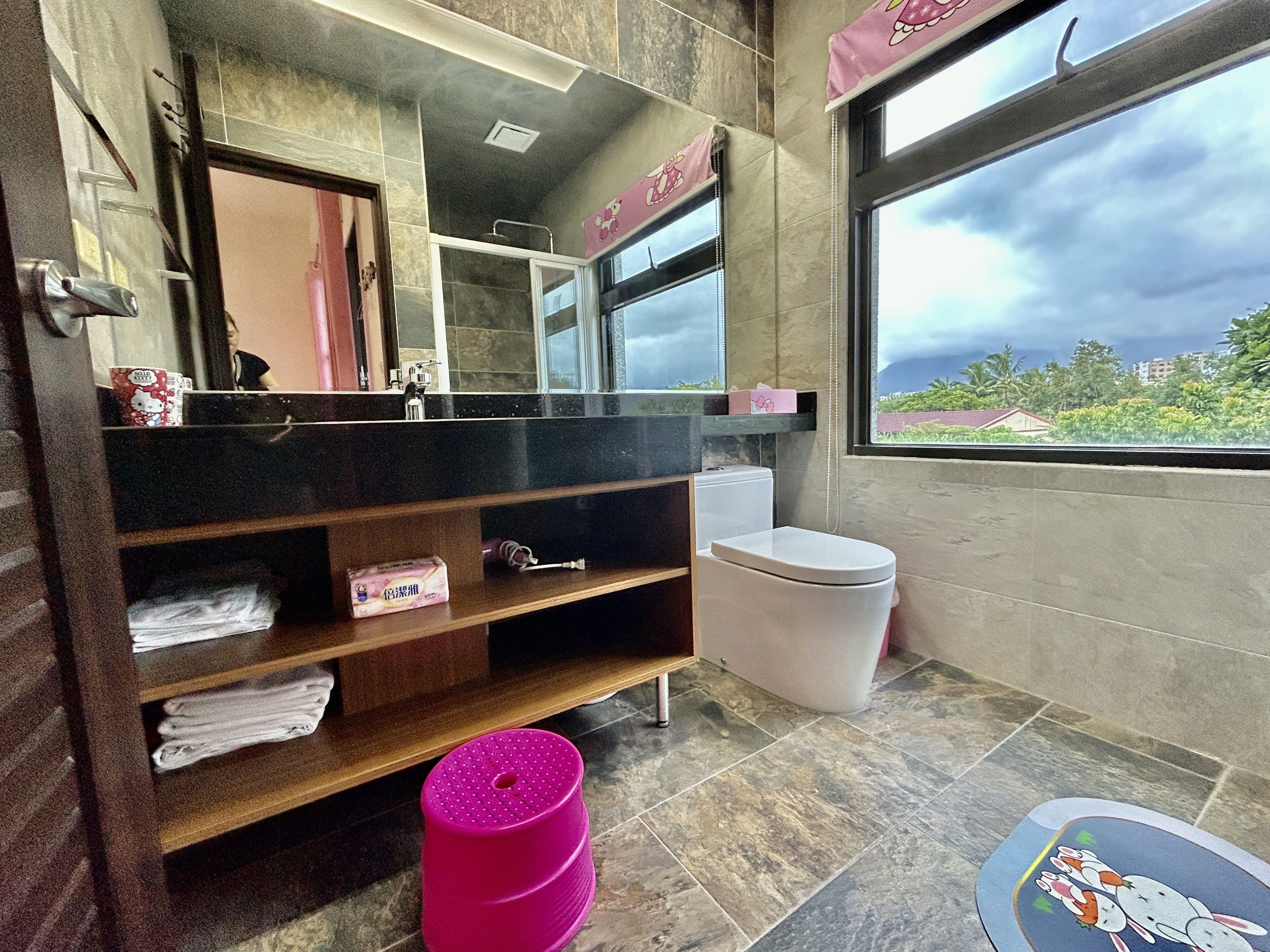 A bathroom with a large mirror, a toilet, and a sink. There is a window with a view of greenery and buildings outside.