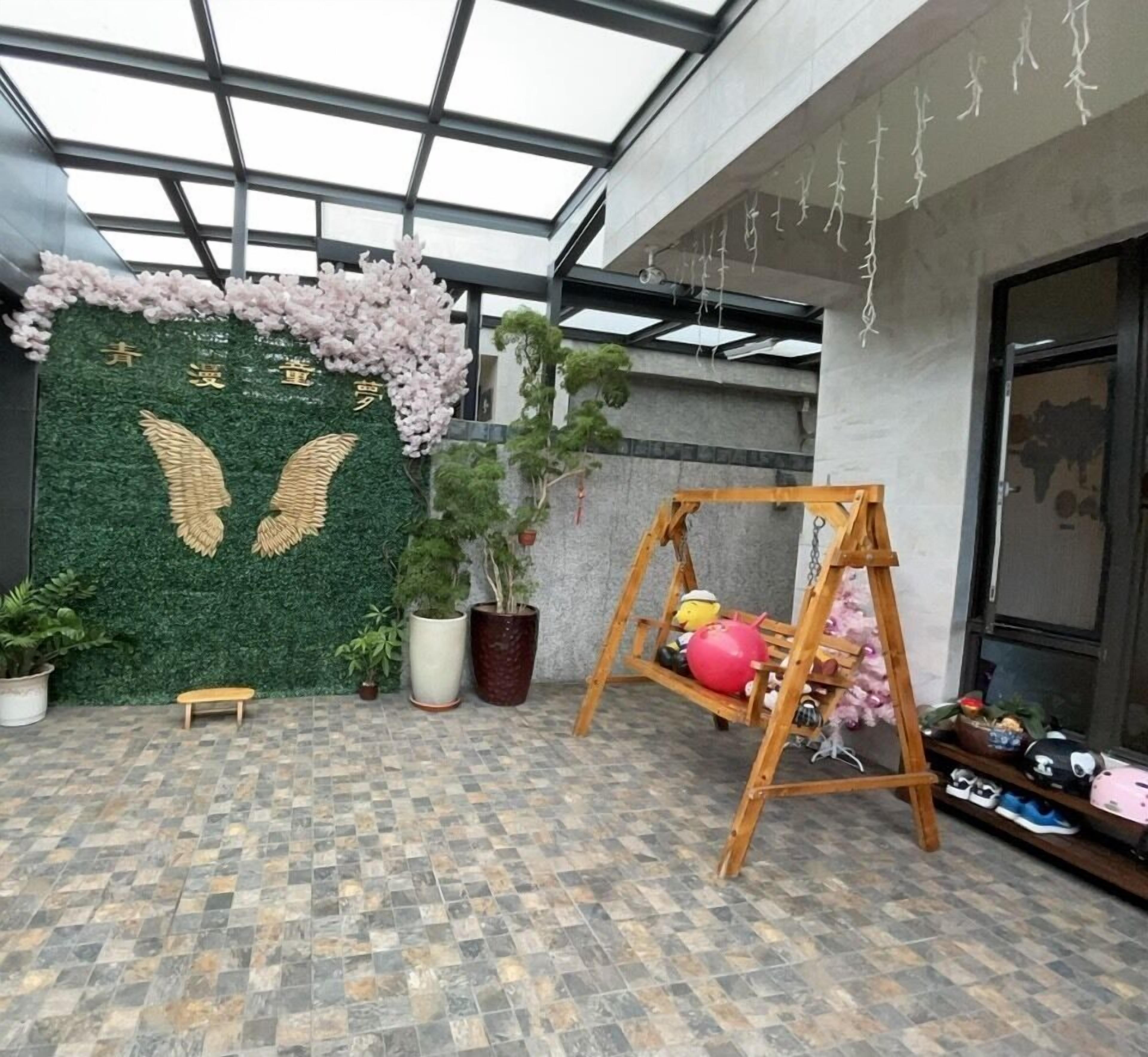 A room with a green wall decorated with golden wings and pink flowers, a swing, and various potted plants.