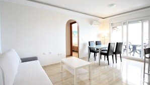 Un salon moderne avec un canapé blanc, une petite table blanche et un coin repas comprenant une table en verre et des chaises noires. Une porte donne accès à un balcon aménagé avec du mobilier d’extérieur.
