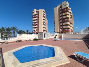 A rooftop pool area with a rectangular pool, lounge chairs, and two tall buildings in the background. - Modern one bedroom only 50m from the beach (La Manga del Mar Menor)