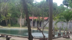 A pool area with a concrete pathway, trees, and a building with a red roof in the background. - Menjangan Hostel (Pejarakan)