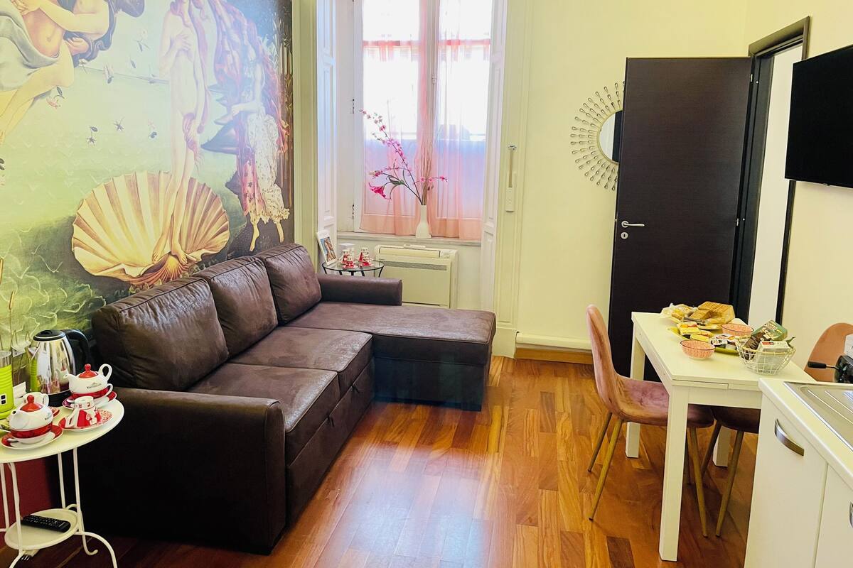 A living room with a brown leather sofa, a small table with a teapot, a window with pink curtains, and a wall-mounted clock.