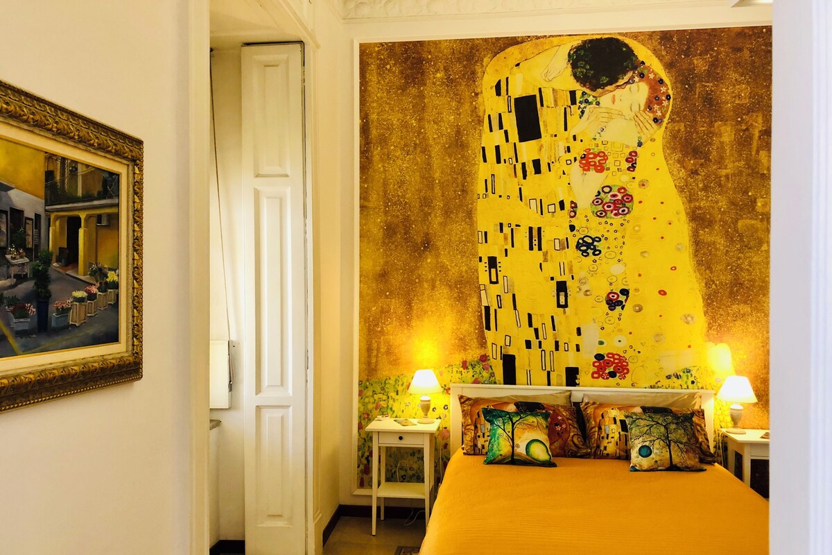 Ett hotellrum med en stor målning av Gustav Klimt, en säng med ett gult överkast, två sängbord med lampor och en dörr som leder till ett annat rum.