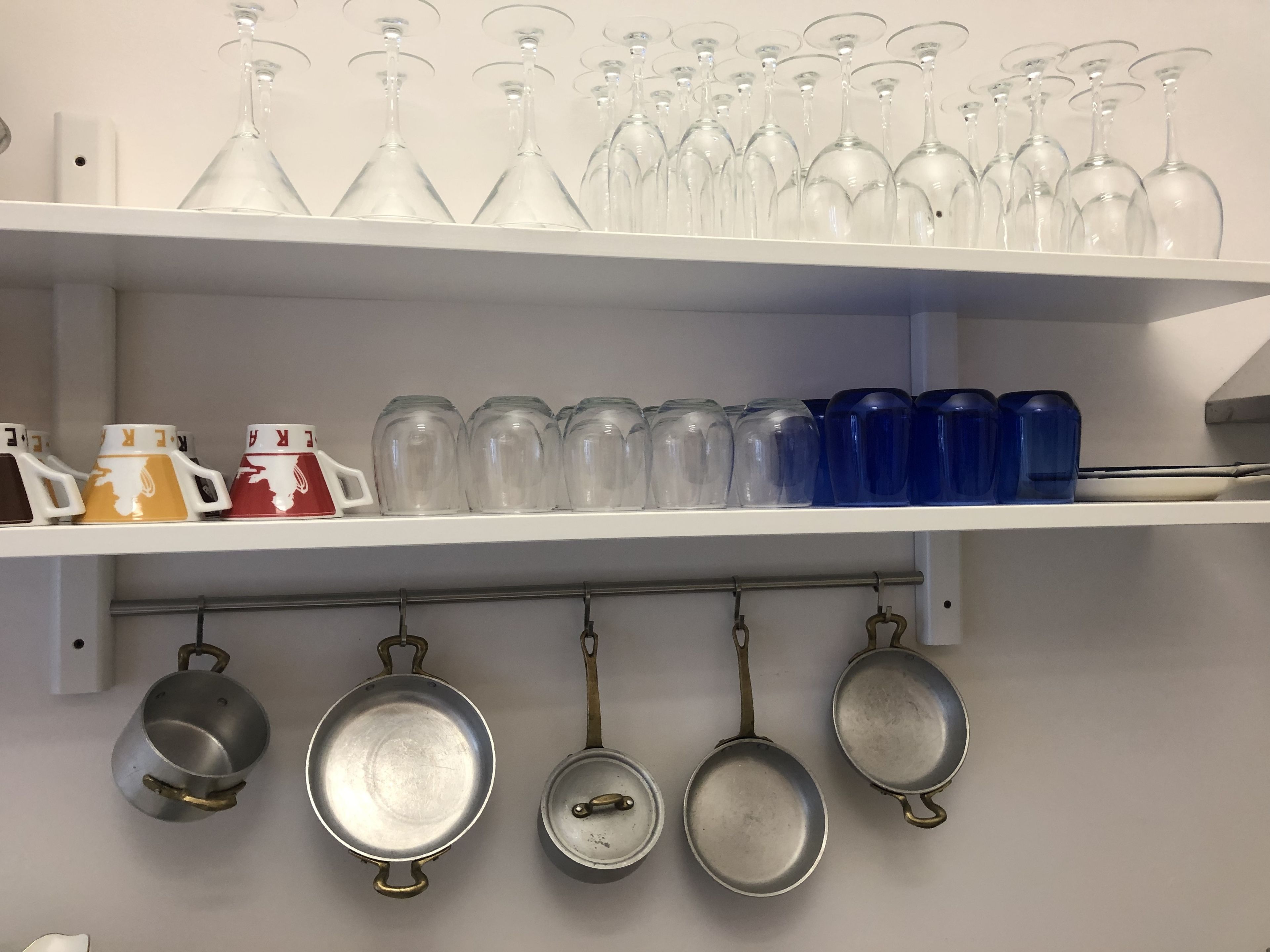 A kitchen with shelves holding wine glasses, jars, and hanging pots.