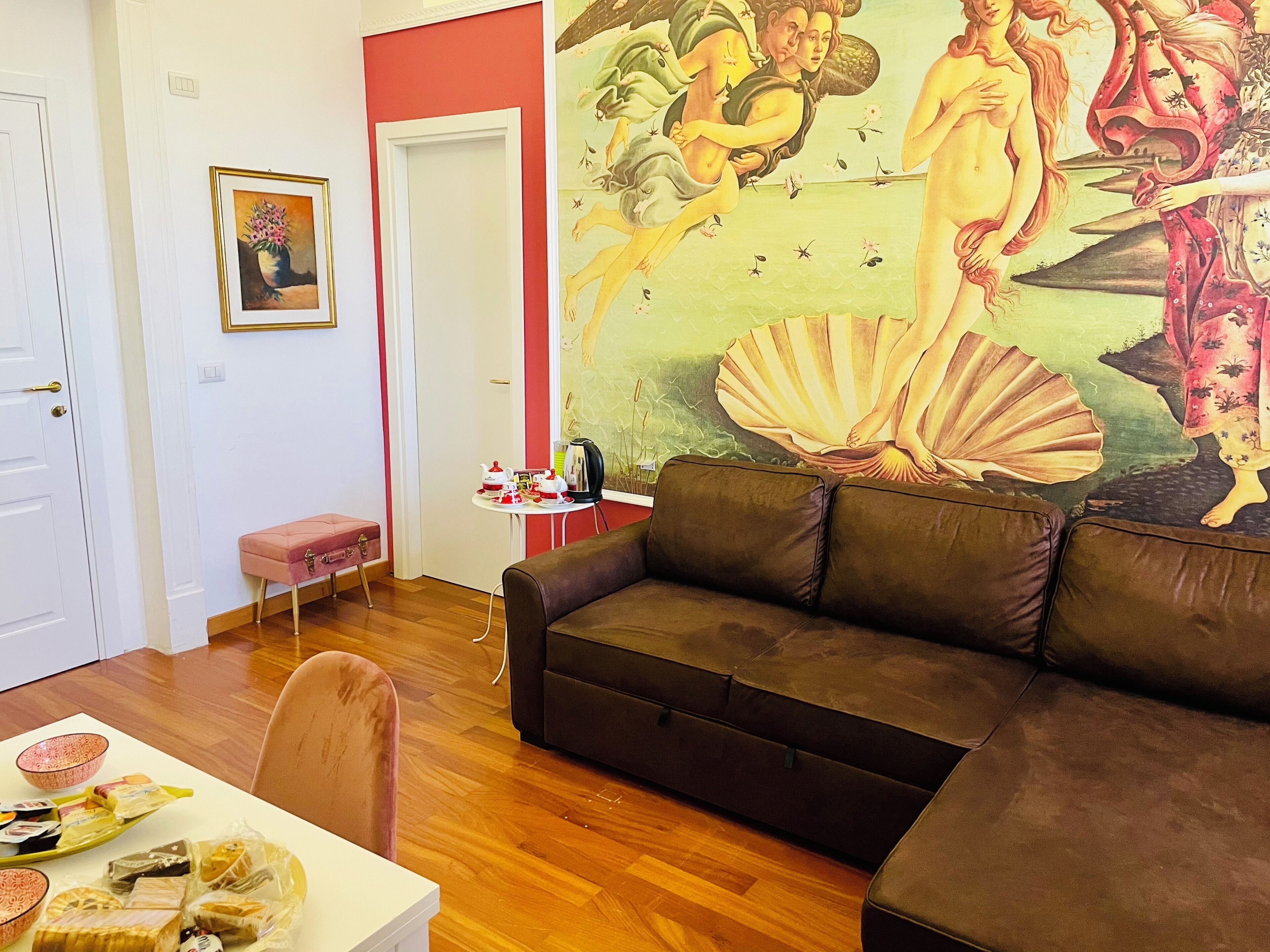 A living room with a brown sofa, a dining table with food, a pink ottoman, and a mural on the wall.