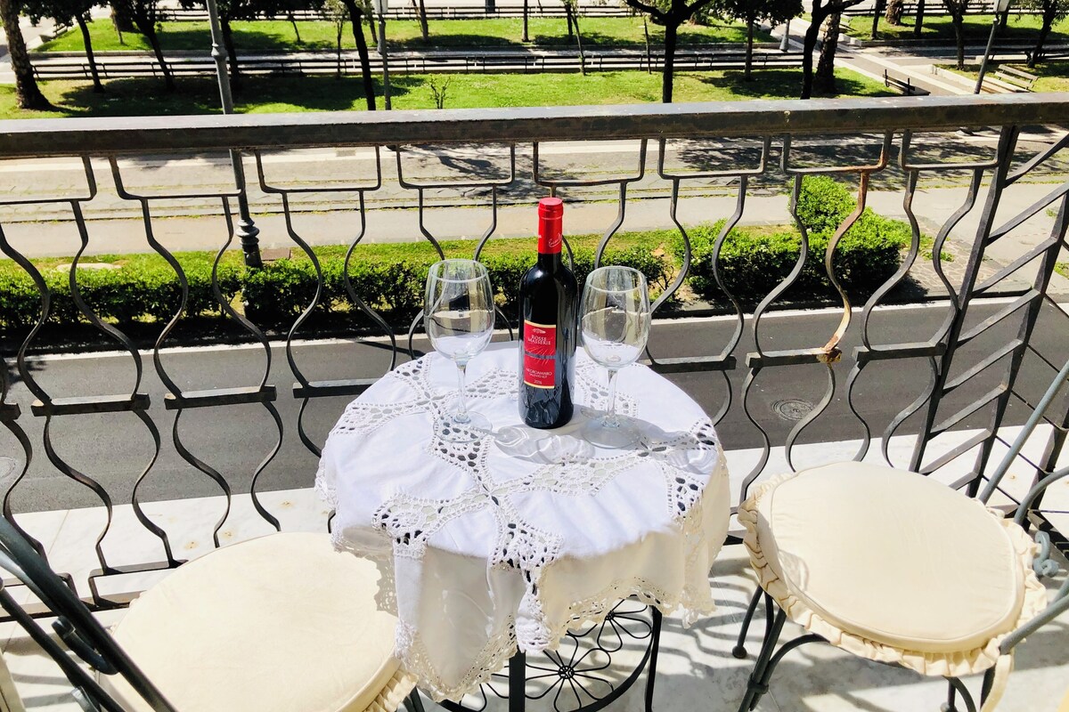 Una mesa con una botella de vino, dos copas y un mantel de encaje en un balcón exterior.