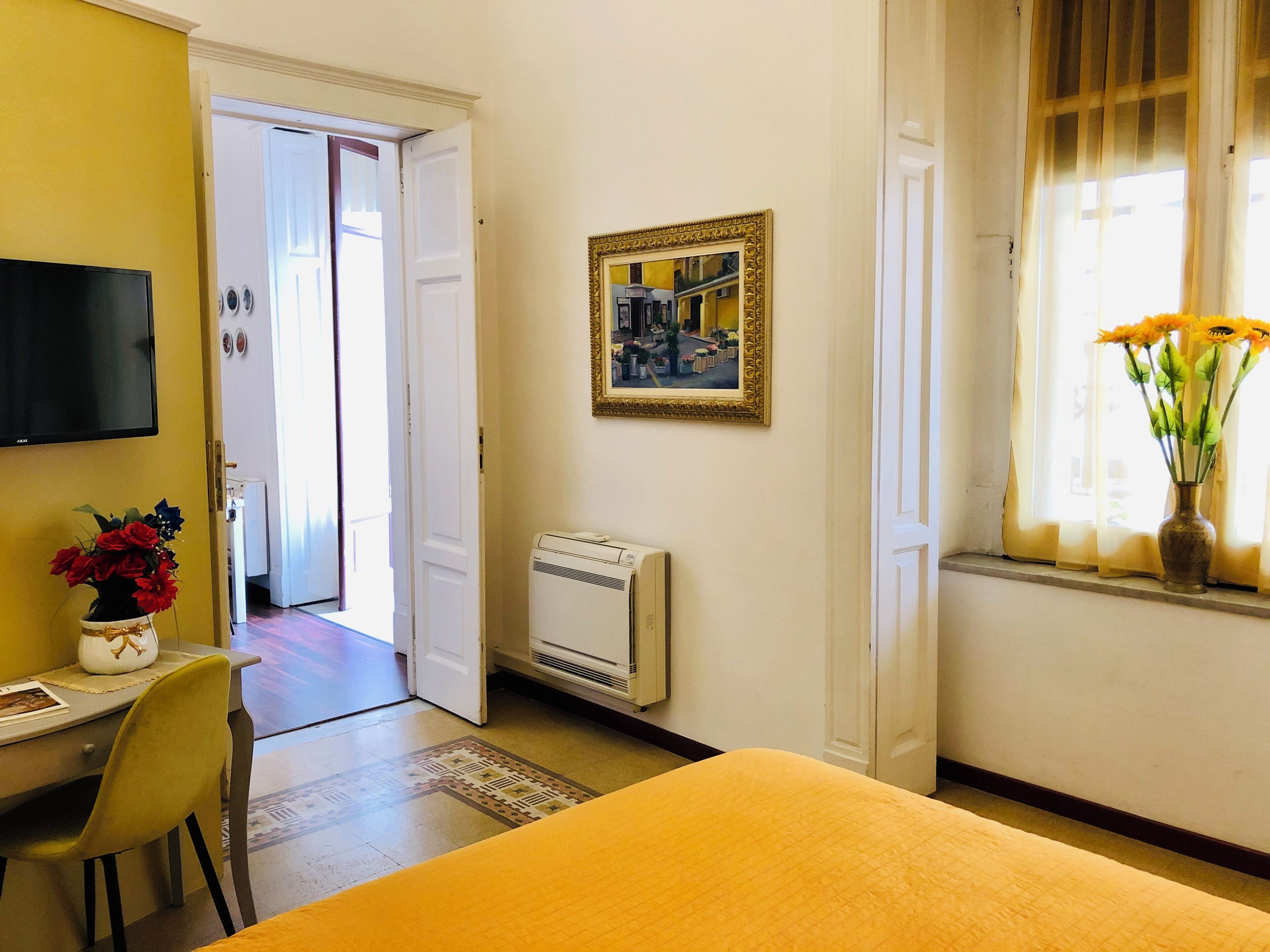 Ein Hotelzimmer mit einem Bett, einem Schreibtisch mit Stuhl, einem Fernseher, einem Heizkörper, einem gerahmten Bild und einem Fenster mit Blumen.