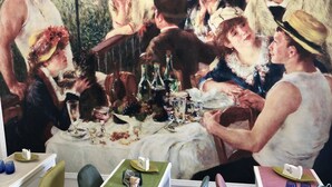 A dining area with a large painting depicting a historical scene, a table set with plates, glasses, and bottles, and chairs with green cushions.