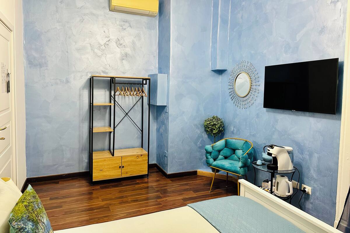 A bedroom with a bed, a wardrobe, a chair, a TV, and a coffee machine.