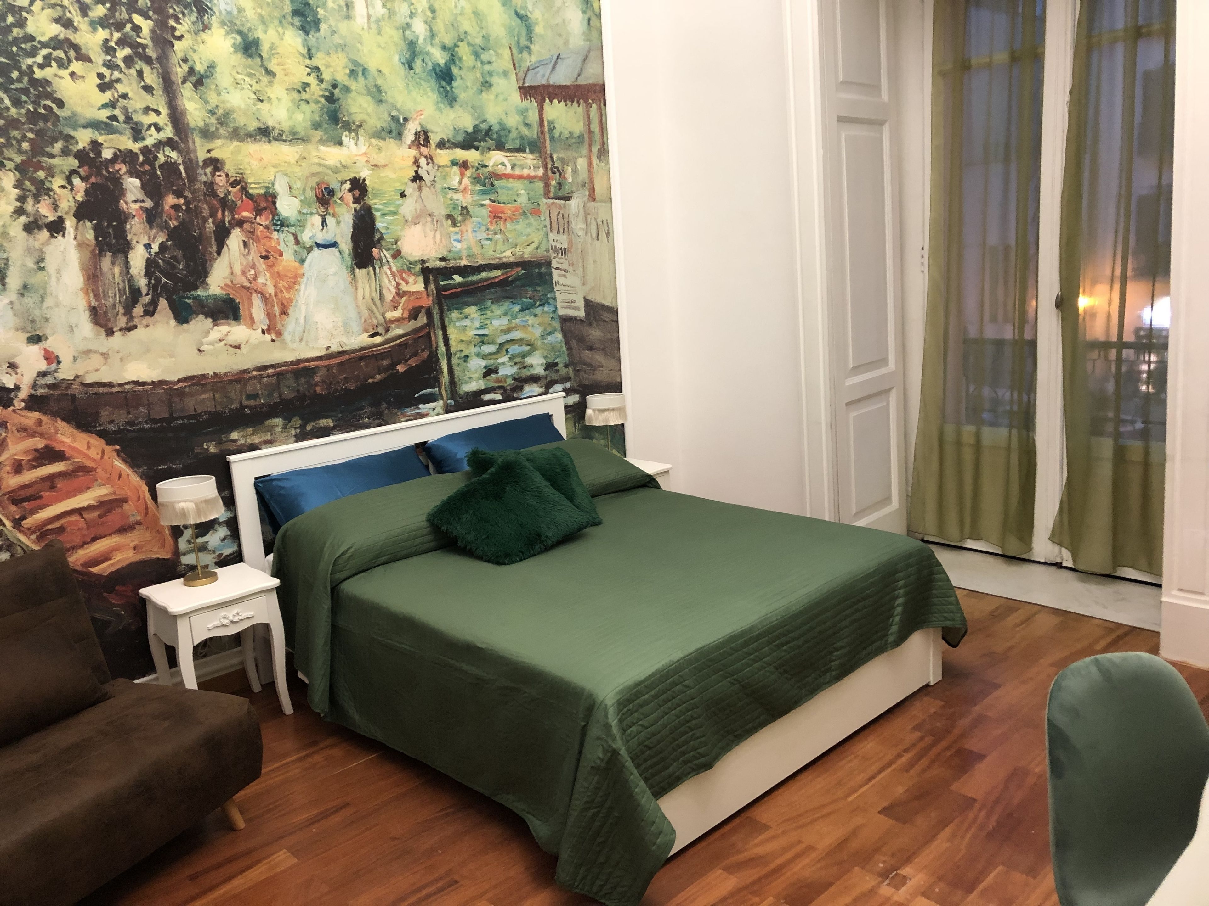 Un dormitorio con un cuadro grande en la pared, una cama con ropa de cama verde, una mesita de noche, una silla y una ventana con cortinas.