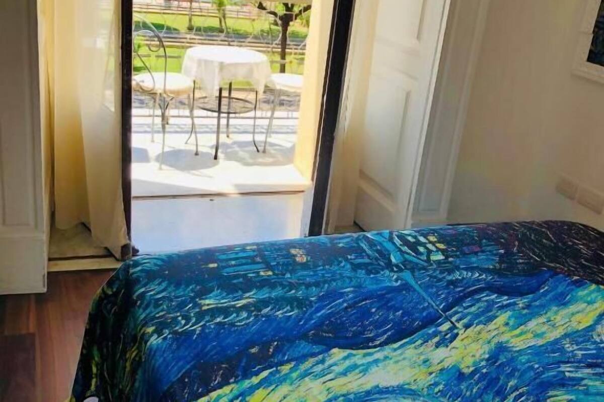 A bedroom with a bed covered in a blue and yellow patterned bedspread, an open door leading to a balcony with a table and chairs, and a view of the sea.