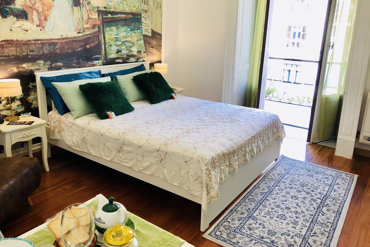 A bedroom with a bed, a nightstand, a painting, a window, and a view of the outside.