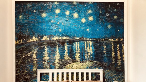 A bedroom with a large painting of a night scene with stars and water, a white bed frame, and two bedside tables with lamps.