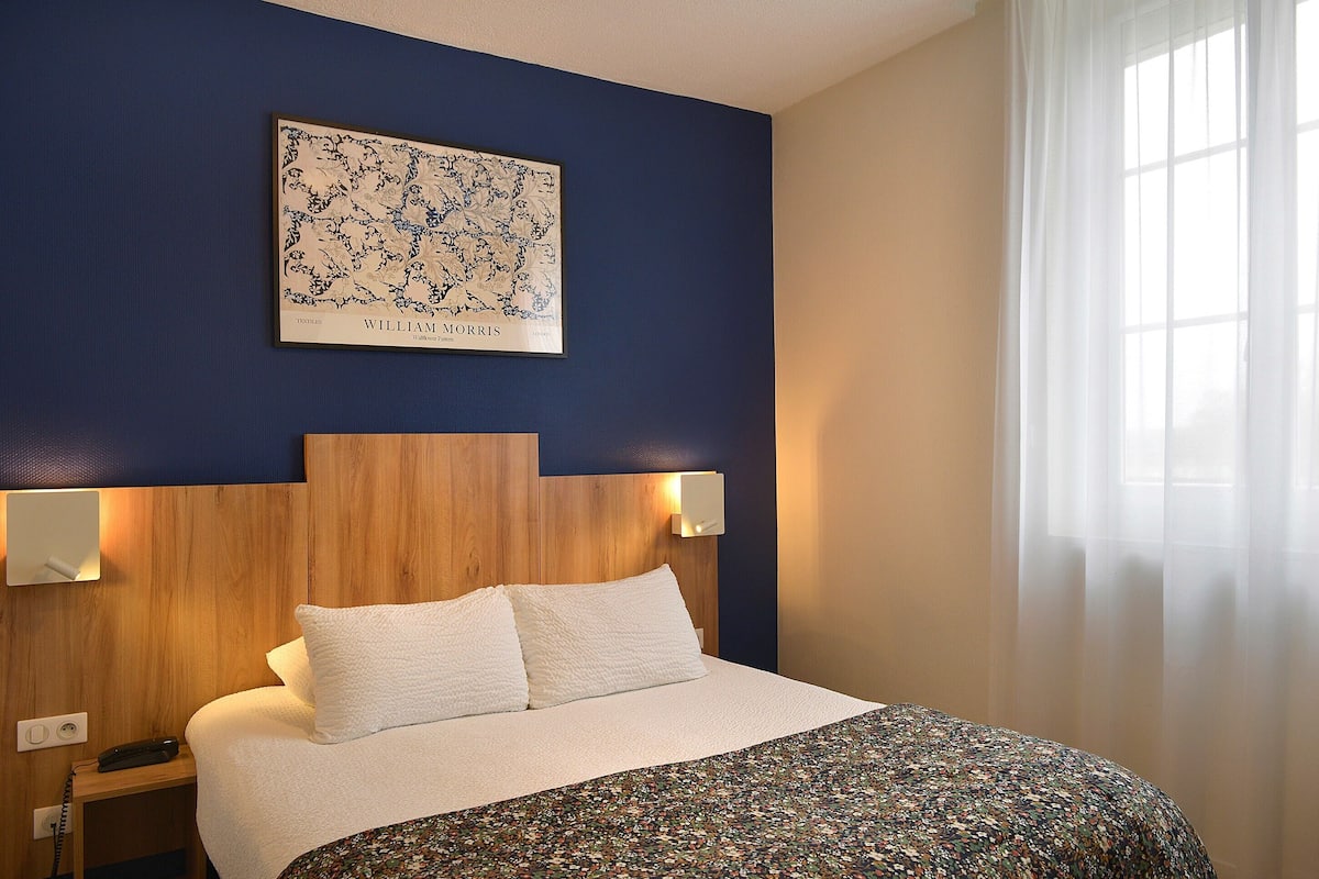 Habitación de hotel con cabecera de madera, ropa de cama blanca, colcha de estampado floral, un mapa enmarcado en la pared y dos lámparas de noche.