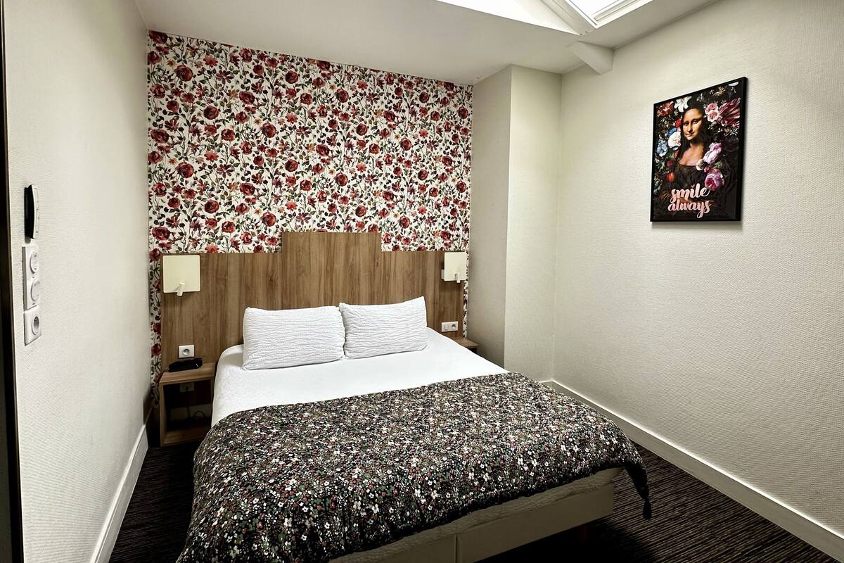 Habitación de hotel con una cama, cabecera de madera, pared con estampado floral, tragaluz y un cartel en la pared.