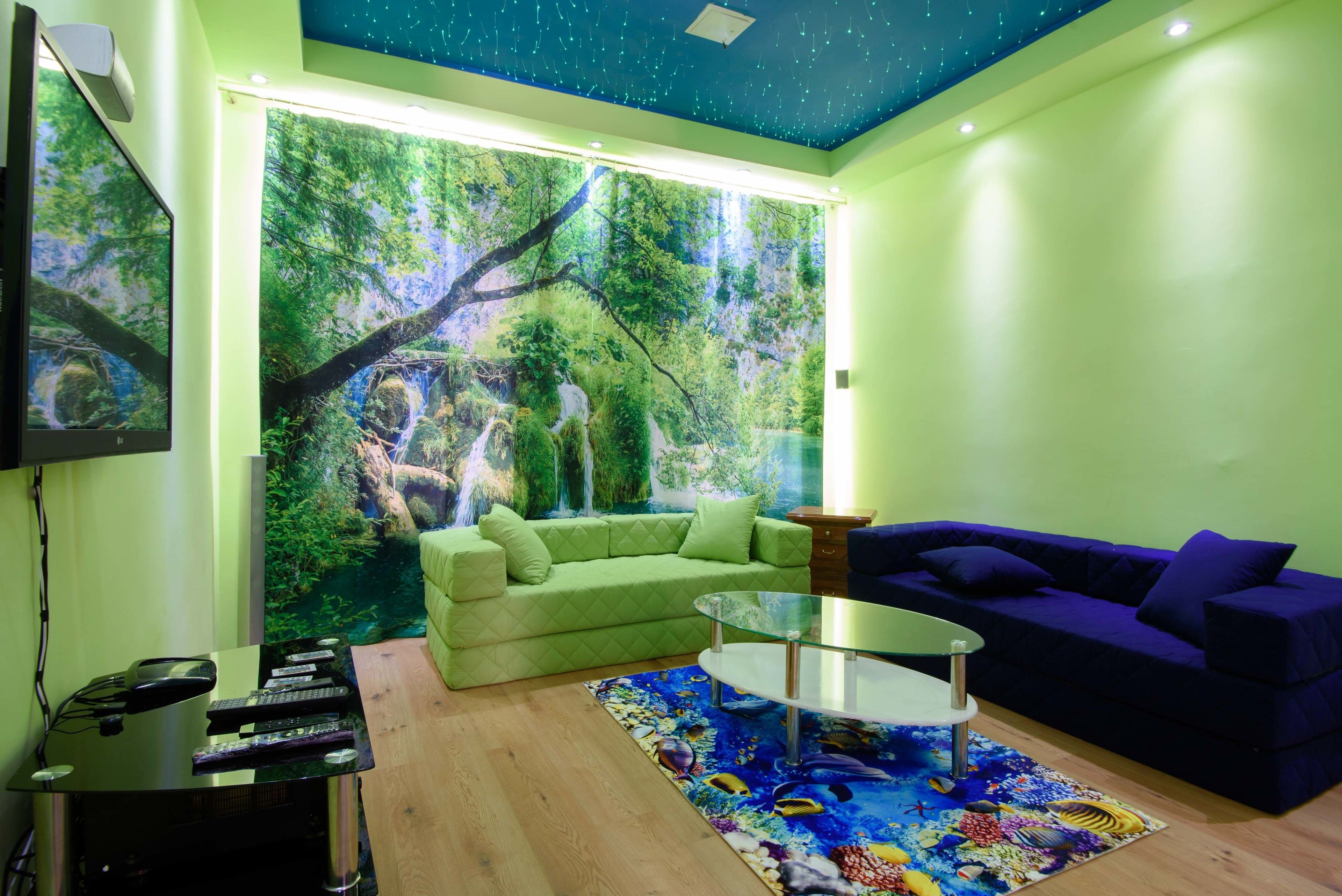 A living room with a green sofa, a blue sofa, a glass coffee table, and a large wall mural of a forest scene.