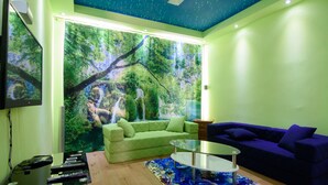 A living room with a green sofa, a blue sofa, a glass coffee table, and a large wall mural of a forest scene.
