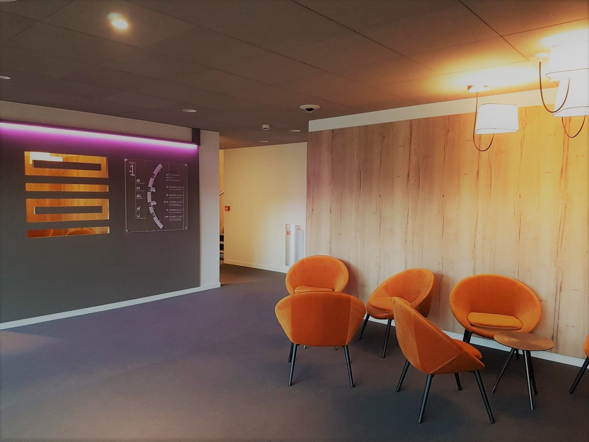 A reception area with a modern design, featuring a reception desk, a wall-mounted information board, and a row of orange chairs.