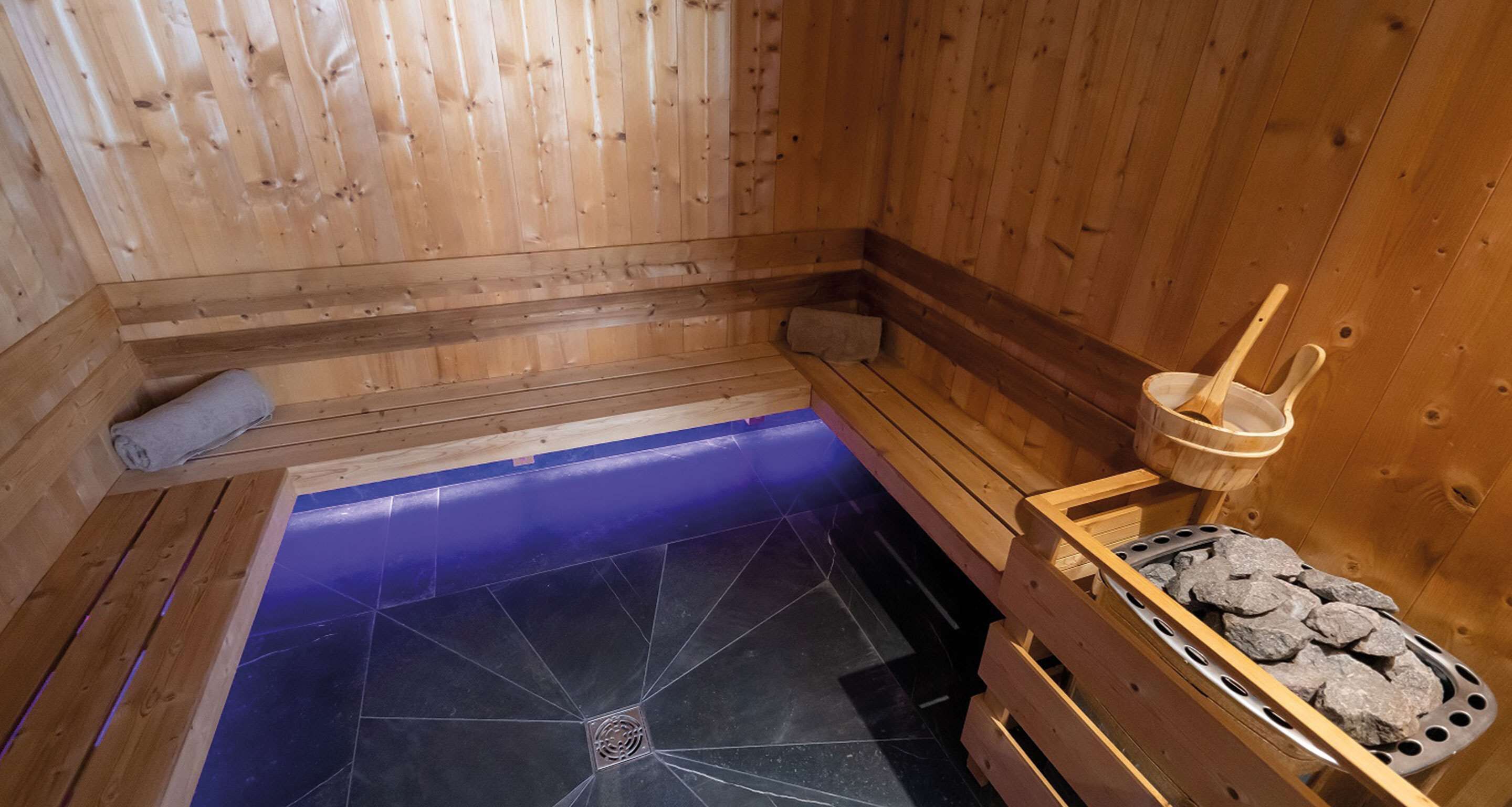 Een houten sauna met een blauw betegelde vloer, houten banken, een handdoek en een emmer met stenen.