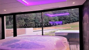 A modern spa room with a large hot tub, large windows offering a view of a garden, and purple ambient lighting. - Mildiss Hotel Spa & Restaurant, BW Signature Collection (Besse-et-Saint-Anastaise)