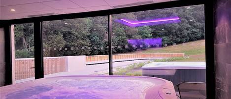 Un spa moderno con una bañera de hidromasaje grande, ventanas amplias que dan a un jardín, e iluminación ambiental morada.