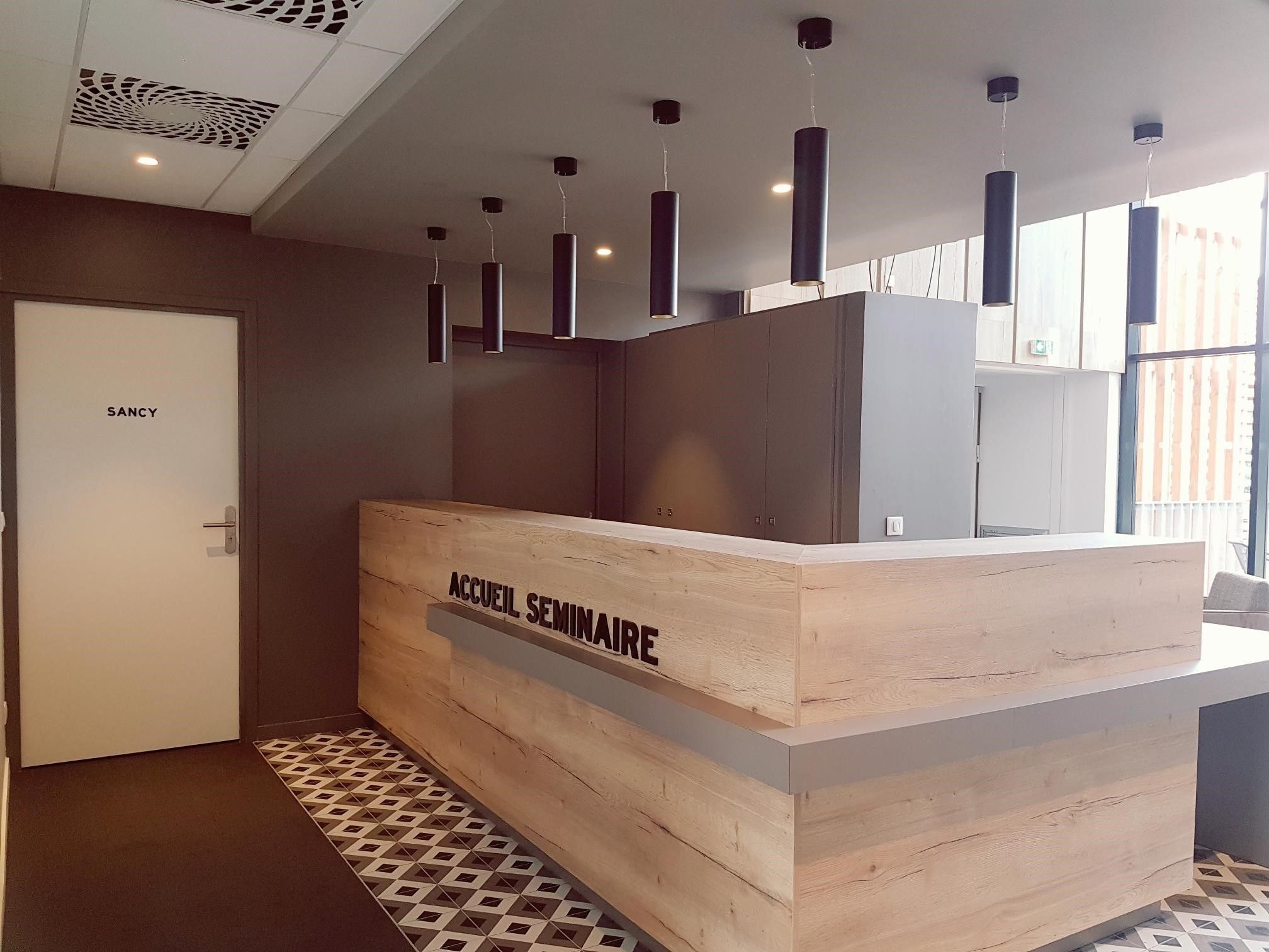 A reception desk labeled "ACCUEIL SEMINAIRE" in a modern hotel lobby with a patterned floor, a door labeled "SANCY", and pendant lighting.