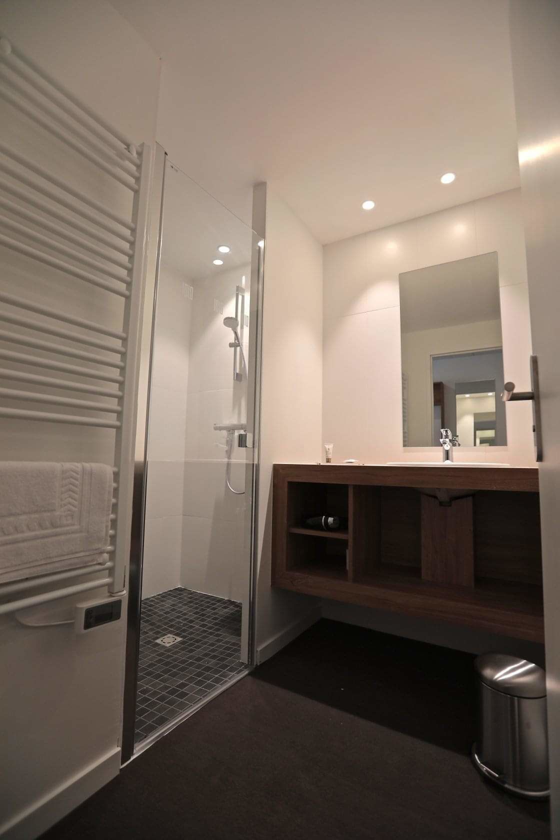 Un baño moderno con ducha de vidrio, tocador de madera con espejo y piso oscuro.