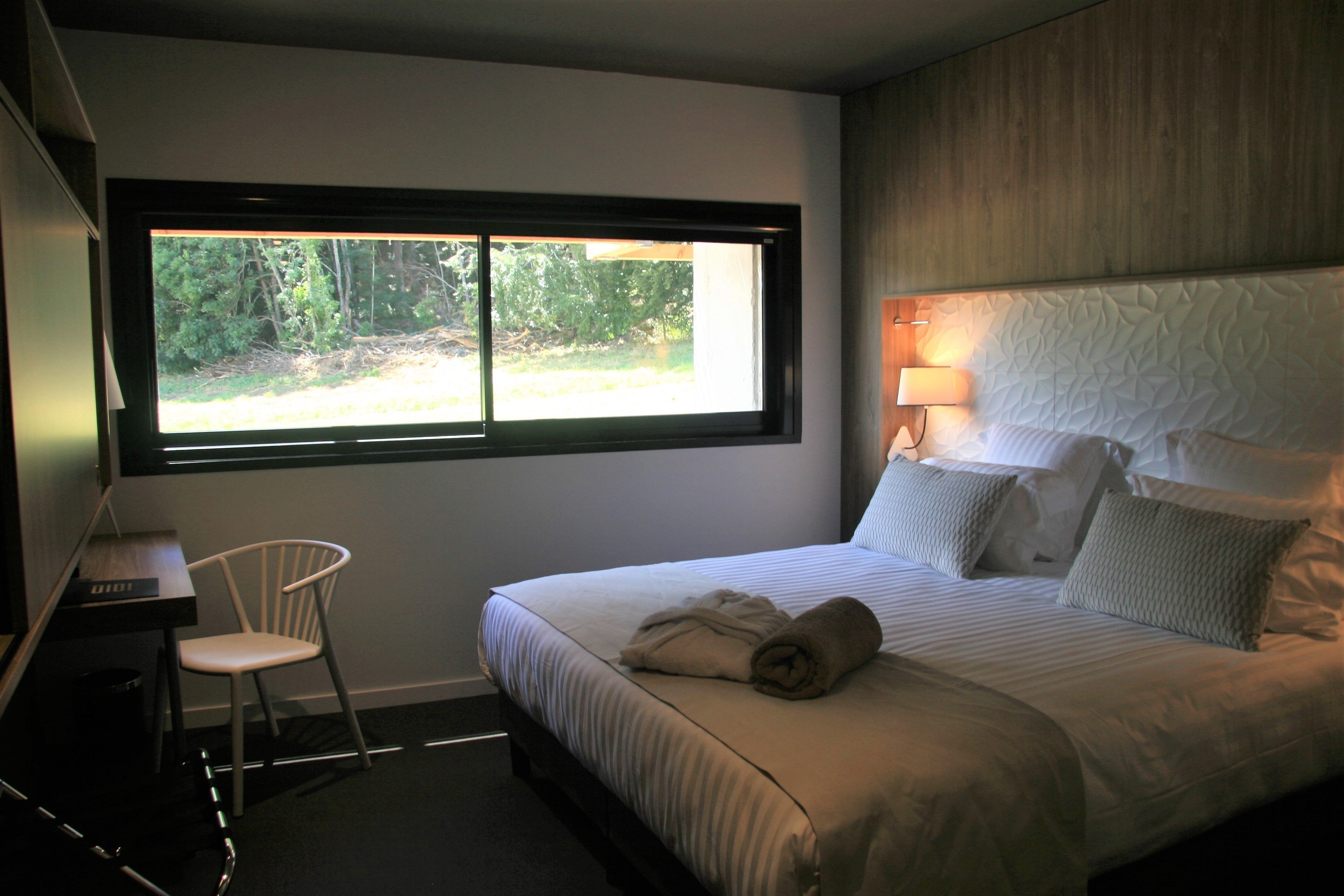 A hotel room with a large bed, a desk with a chair, a window with a view of trees, and a wall-mounted lamp.