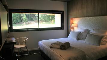 A hotel room with a large bed, a desk with a chair, a window with a view of trees, and a wall-mounted lamp.