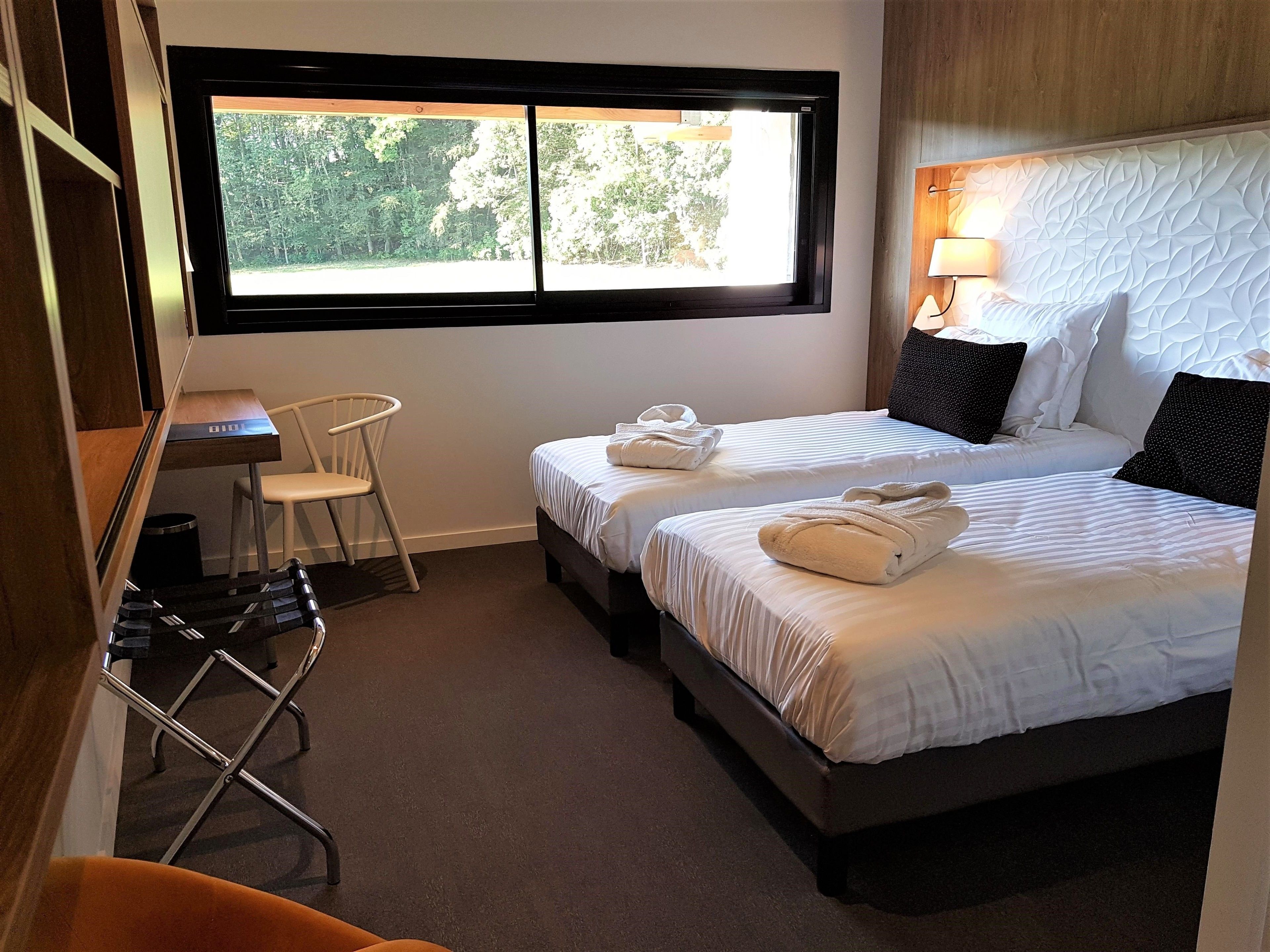 A hotel room with two beds, a desk, a chair, and a window with a view of trees.