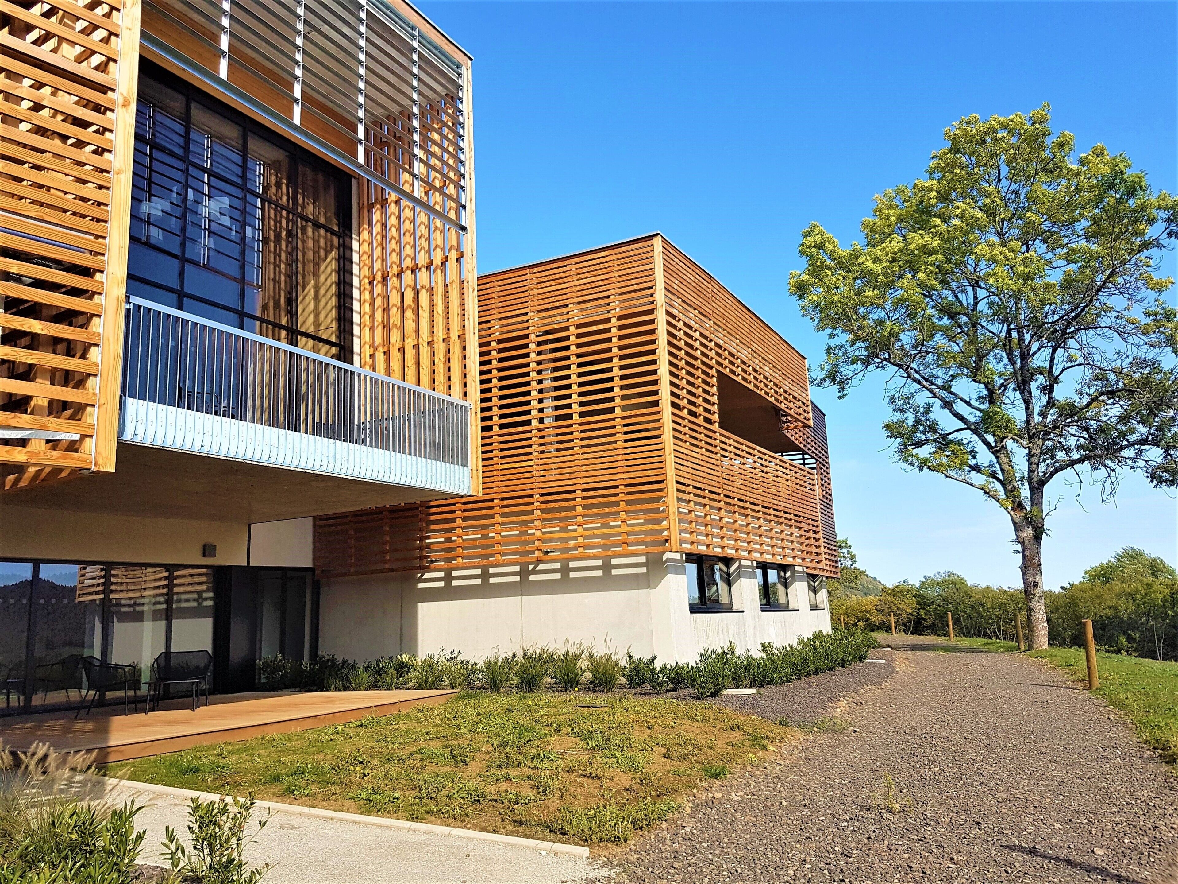 A modern building with a wooden facade, a balcony, and a gravel pathway leading to a tree.